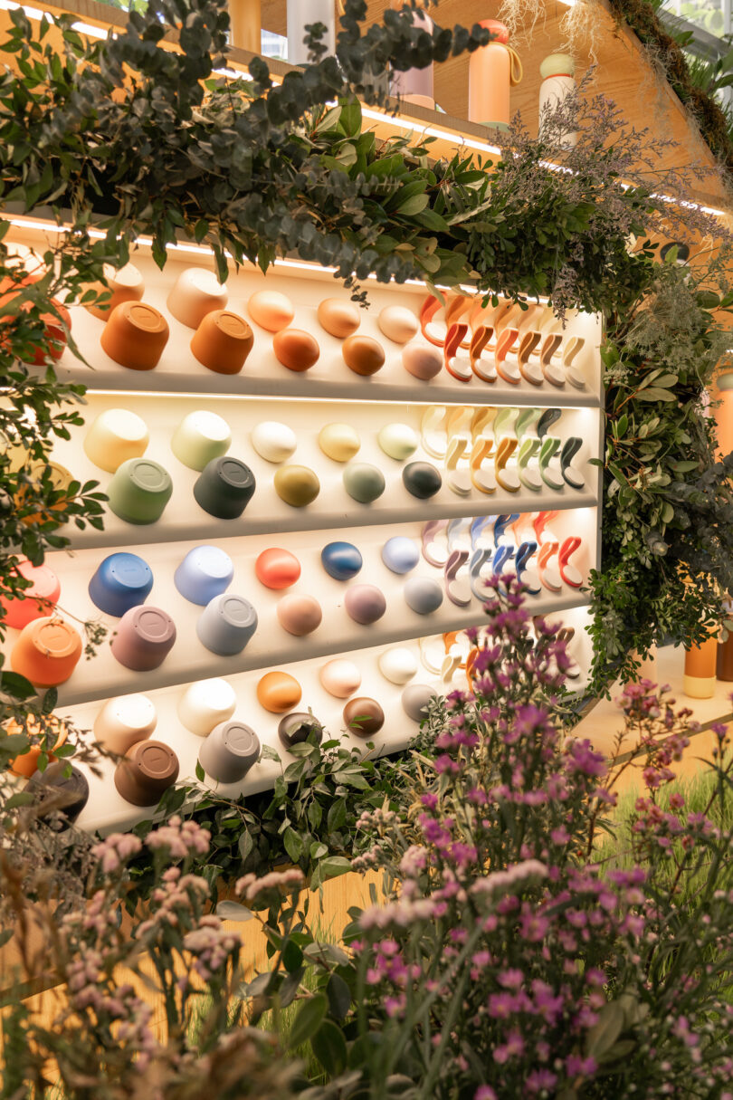 A display wall presenting various colorful coffee mugs, arranged in rows and surrounded by green foliage and flowers.