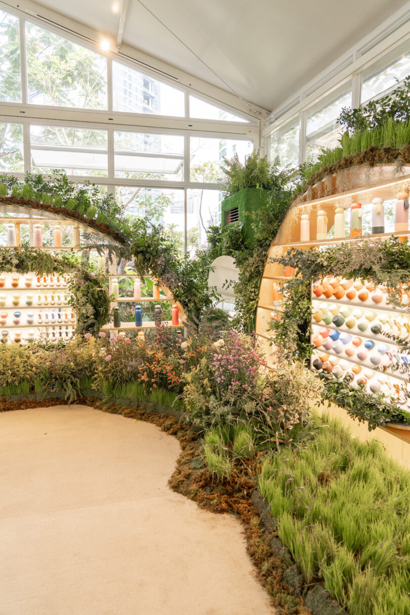 A botanical-style shop with glass walls displaying various bottles and spheres surrounded by lush greenery and plants.