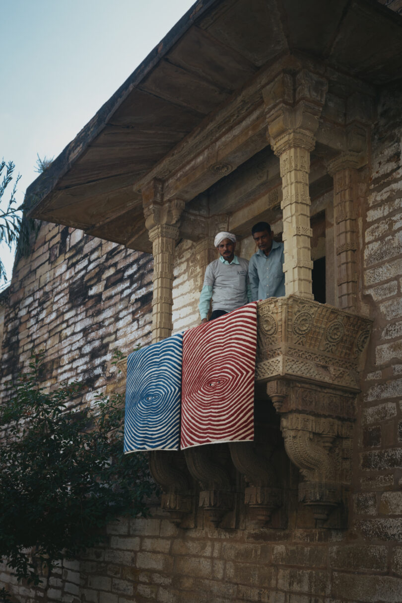 two men standing next to a blue and a red rug hung on a balcony of a brick building