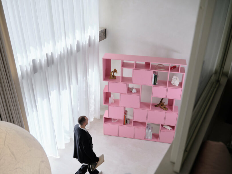 man walking towards a modular pink shelf in a room next to a curtained window