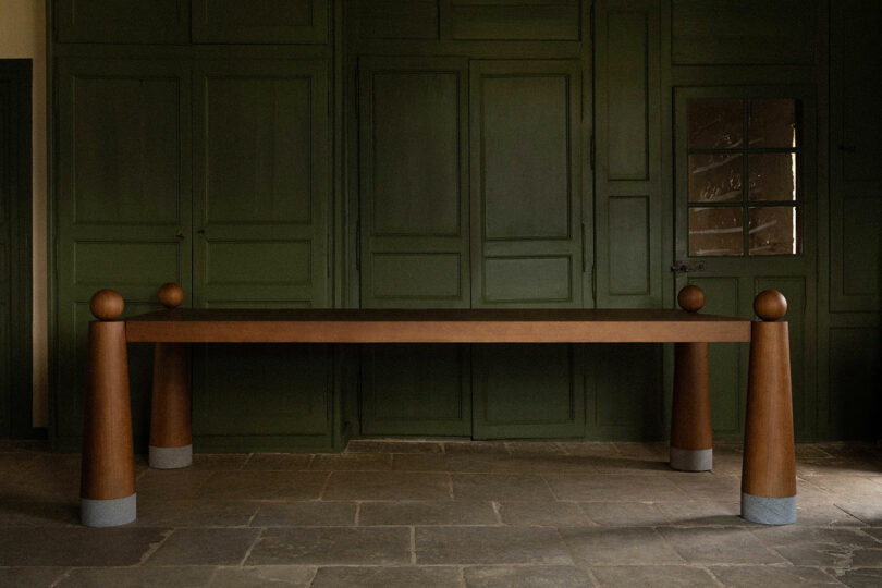 Uma longa mesa de madeira com grossas pernas cilíndricas numa sala com paredes revestidas de verde escuro e chão de pedra.