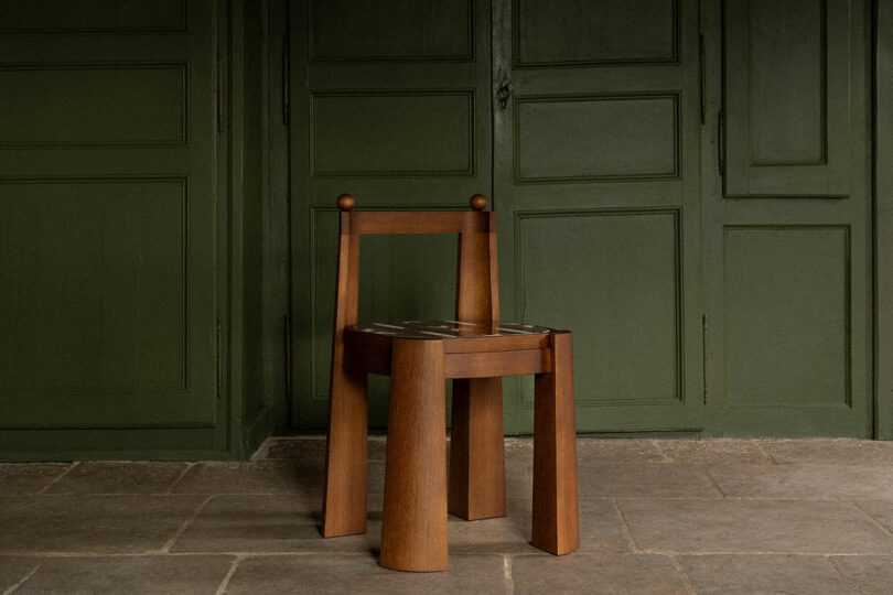 A simple wooden chair stands in front of a green paneled wall on a stone floor.