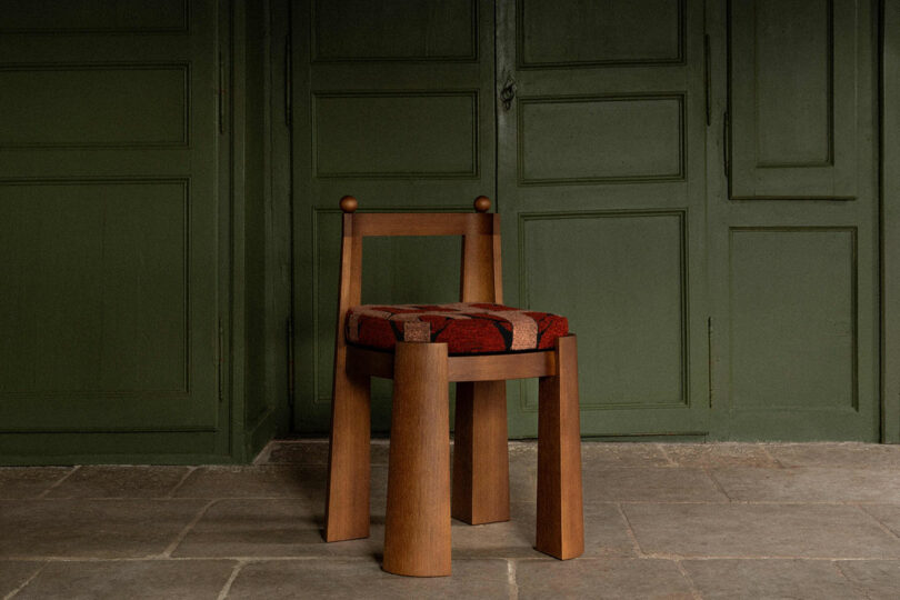 Uma cadeira simples de madeira almofadada fica em frente a uma parede com painéis verdes sobre um chão de pedra.