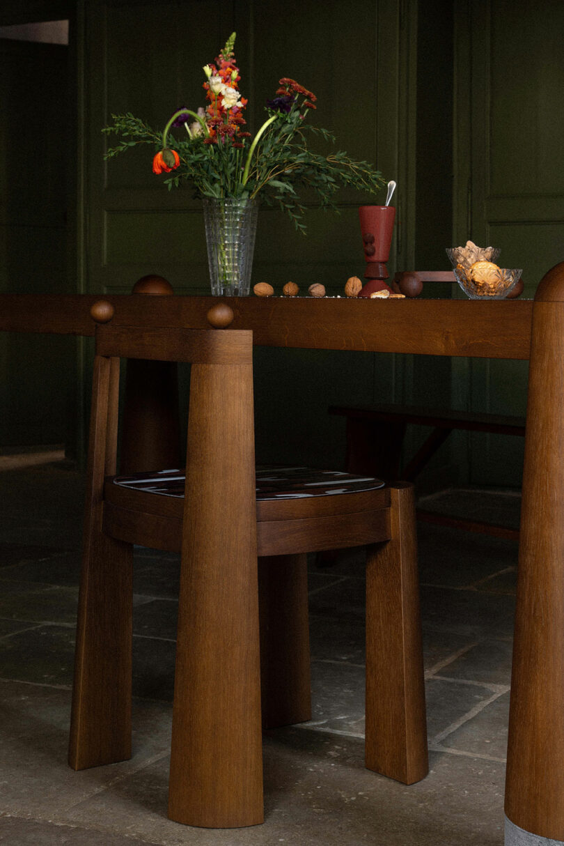 Uma mesa de jantar de madeira com um vaso de flores, uma jarra e uma tigela de nozes em uma sala com paredes verdes e piso de ardósia.