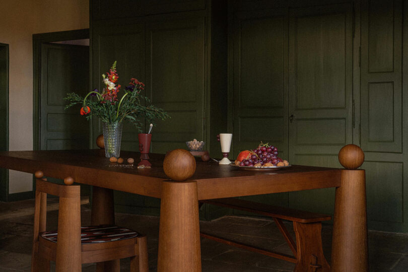 Uma mesa de jantar de madeira com um vaso de flores, uma jarra e uma tigela de nozes em uma sala com paredes verdes e piso de ardósia.