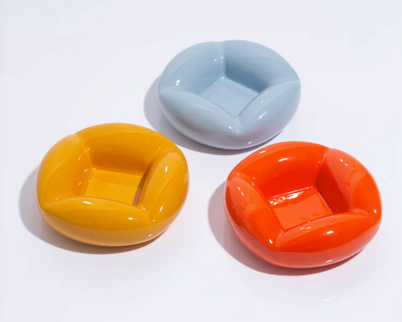 Three colorful ceramic ashtrays in blue, yellow, and orange, artistically arranged on a light background.