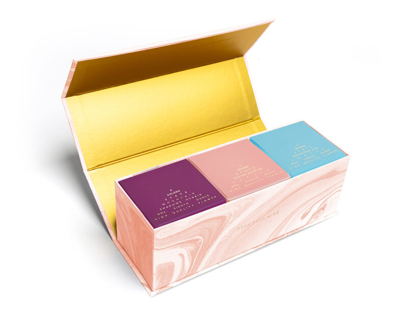 A decorative box opened to reveal three colorful boxes of eighths of cannabis aligned side by side, each featuring a unique pastel cover with gold lettering, including a detailed guide on cannabis cultivation.
