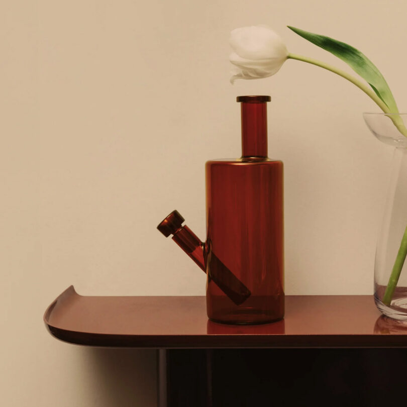 A burnt umber glass water pipe with a white flower on a wooden shelf, next to a vase holding a single tulip.