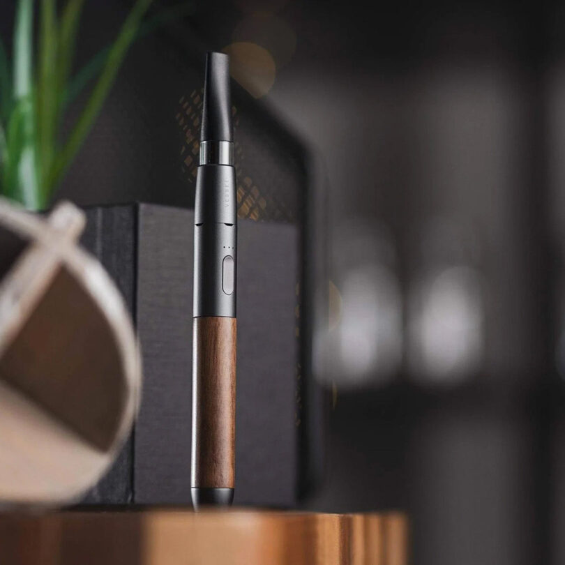 A modern cannabis vaping device with a wooden finish standing upright on a wooden surface, with blurred shelves in the background.