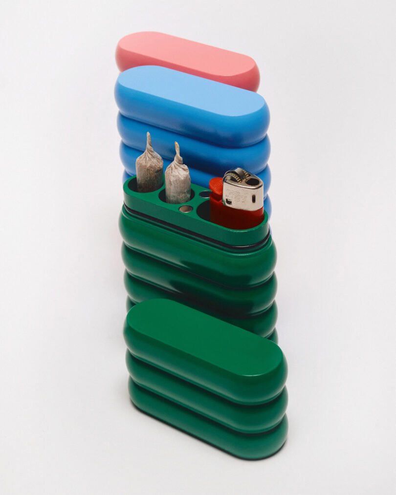 A trio of cannabis stash cases in blue, green, and red, with the green case open showing two joints and red lighter housed within.