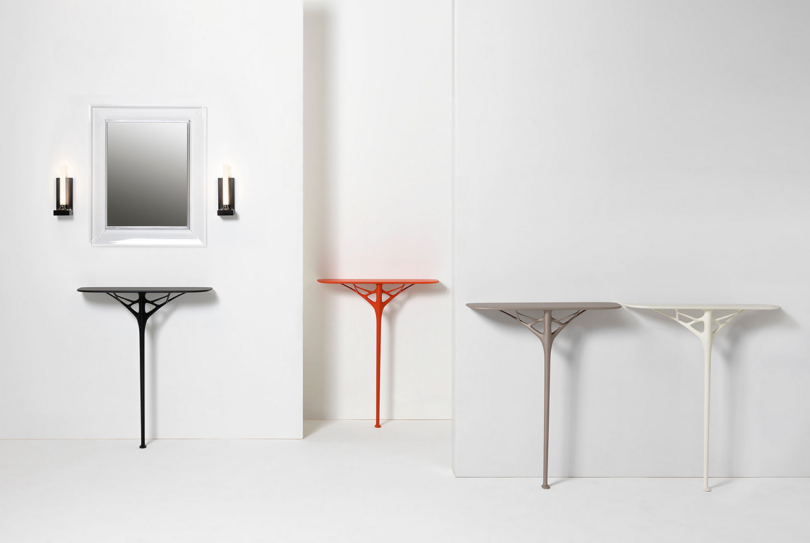 Four minimalist wall consoles in grey, black, orange, and white, placed against a white wall with a framed mirror and wall sconces above.