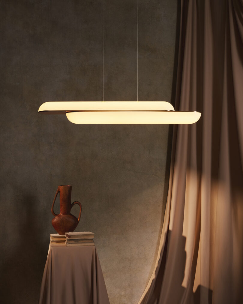 Modern pendant light illuminating a rustic jug on a pedestal, with draped curtains in a dimly lit room with textured walls.