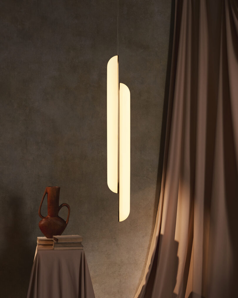 Luminária pendente moderna iluminando um jarro rústico sobre um pedestal, com cortinas drapeadas em uma sala mal iluminada com paredes texturizadas.