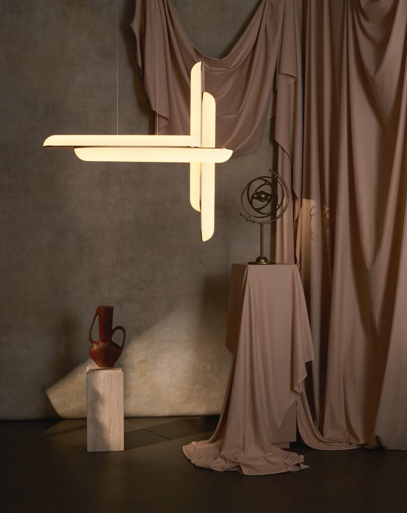 Instalação de iluminação moderna em uma sala com cortinas drapeadas bege, uma escultura de madeira e um ornamento abstrato de metal.