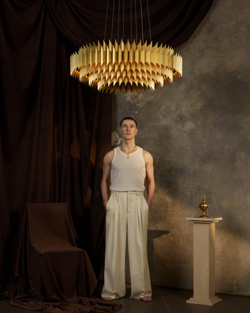 A large, intricate, gold-chandelier hanging against a backdrop of dark draped curtain. A man stands underneath it.