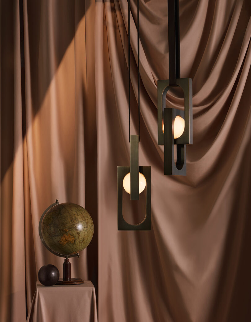 Duas modernas luminárias suspensas com molduras geométricas lançam um brilho quente ao lado de um globo sobre um fundo de tecido drapeado.