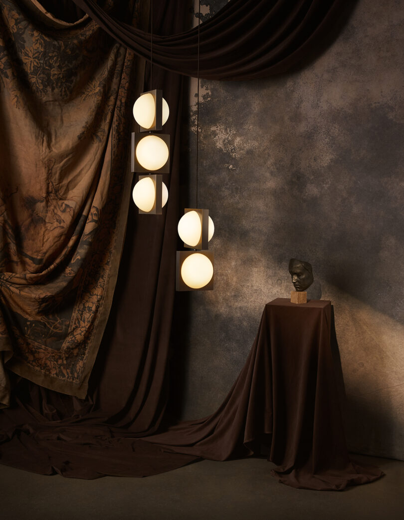 Um interior aconchegante e temperamental com uma luminária suspensa com lâmpadas redondas brilhantes, um busto em um pedestal drapeado e cortinas em uma parede texturizada.