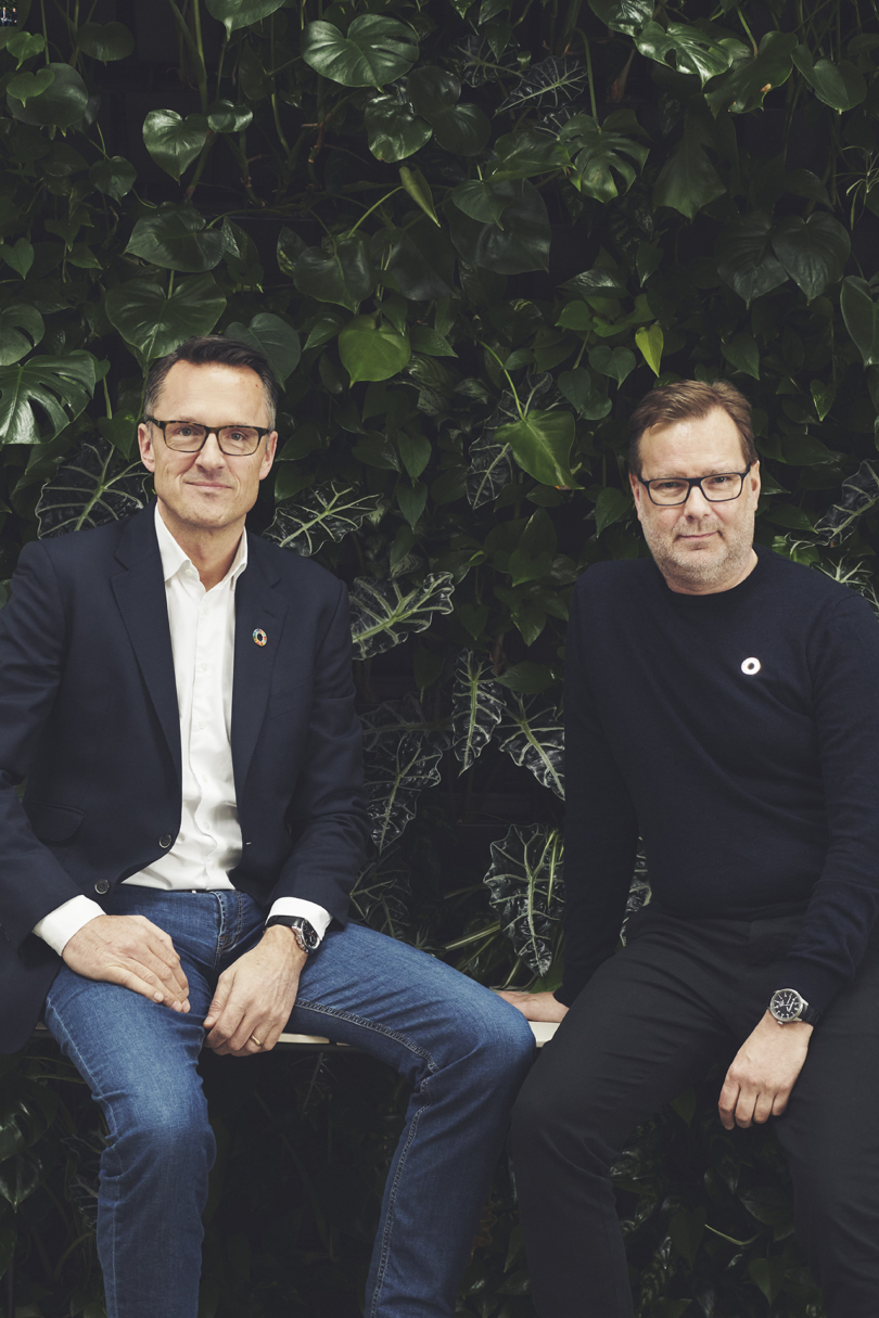 Two white men in casual business attire sitting in front of a leafy green backdrop.