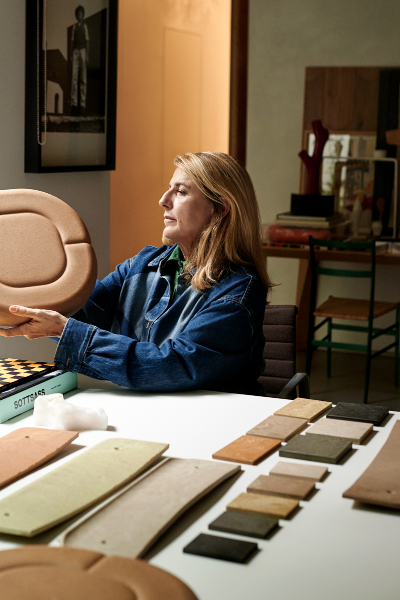 A woman holding a piece of press-molded furniture over a studio table with other materials.