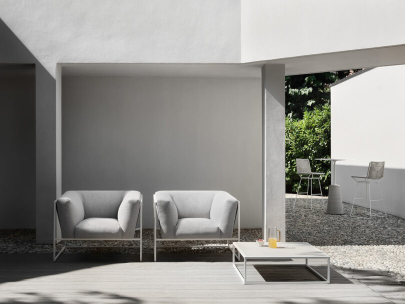 Modern outdoor patio with two armchairs and a coffee table, under a minimalist white architectural structure, surrounded by pebble ground.