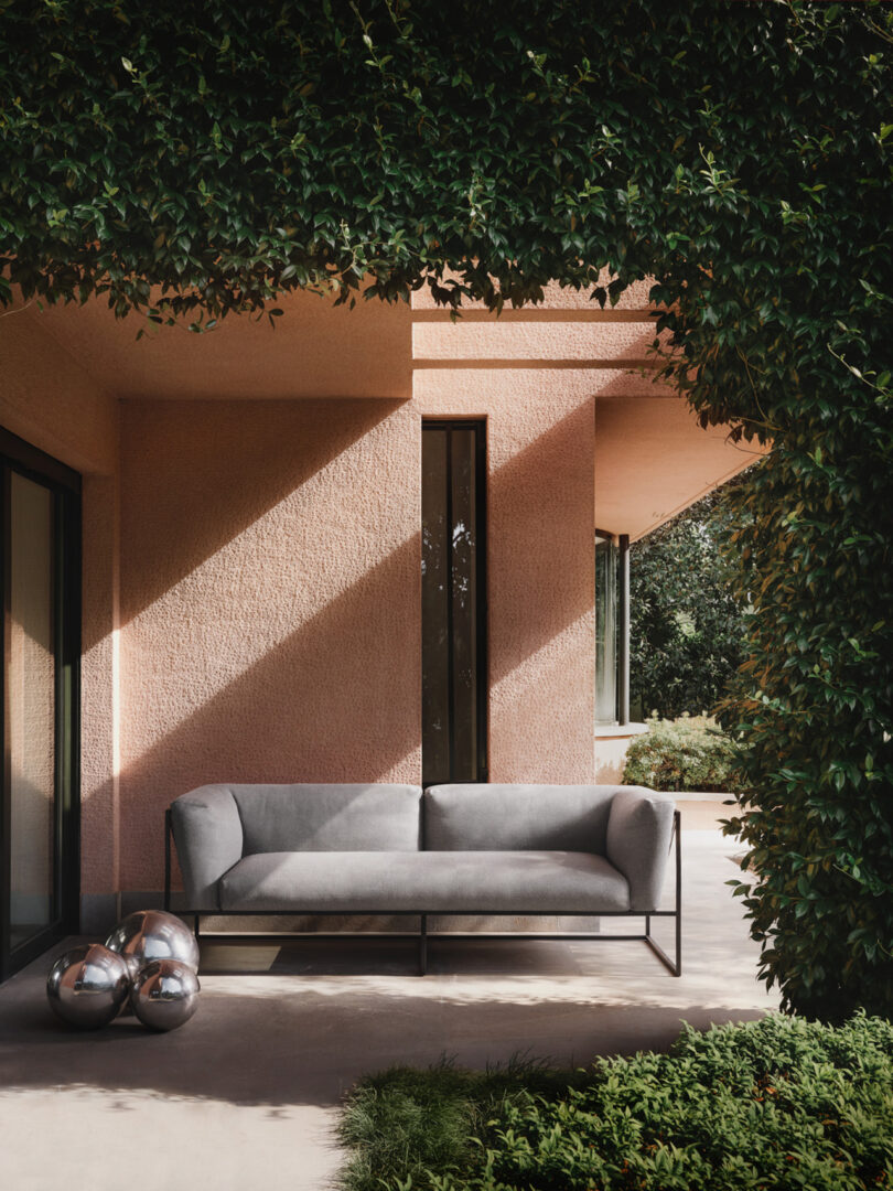 An outdoor seating area featuring a modern gray sofa under the shade of lush greenery, against a textured peach-colored wall with a tall, narrow window.