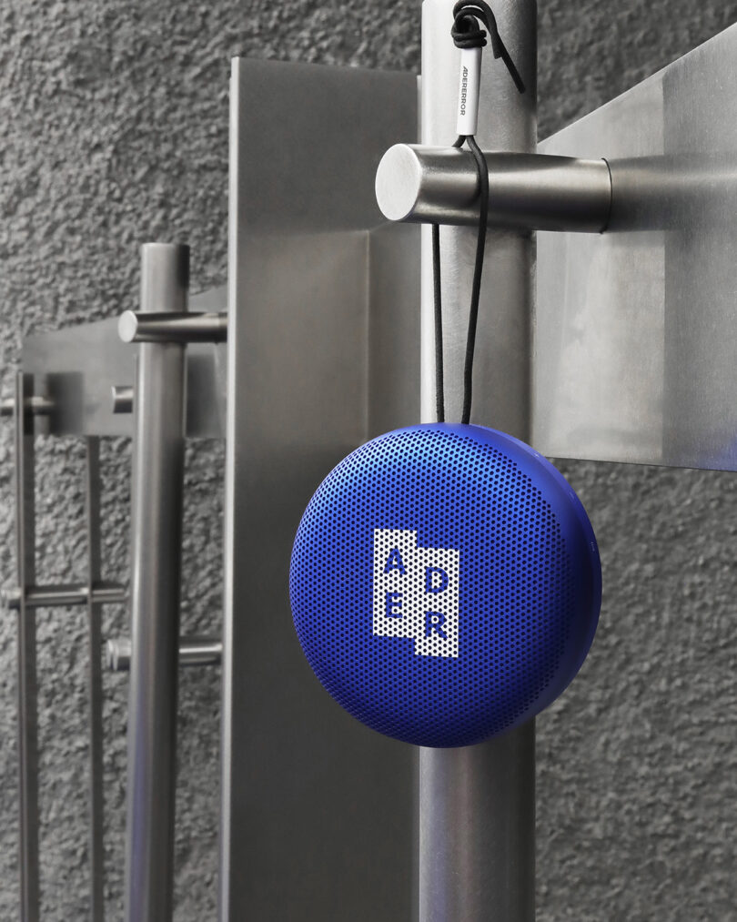 Blue spherical Bluetooth speaker with a perforated front printed with the white logo "Ader", hanging attached to a metal post.