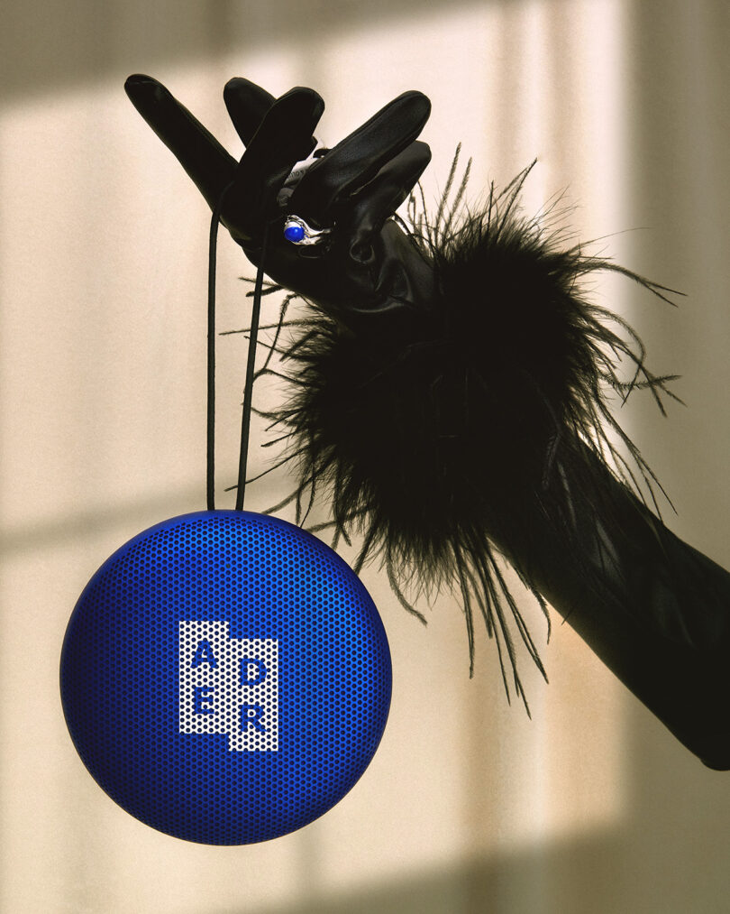 A blue Bang & Olufsen's Beosound A1 ADER ERROR Edition Bluetooth speaker being held by one black gloved hand with a matching blue ring and frilled-fuzzy wrist detailing.