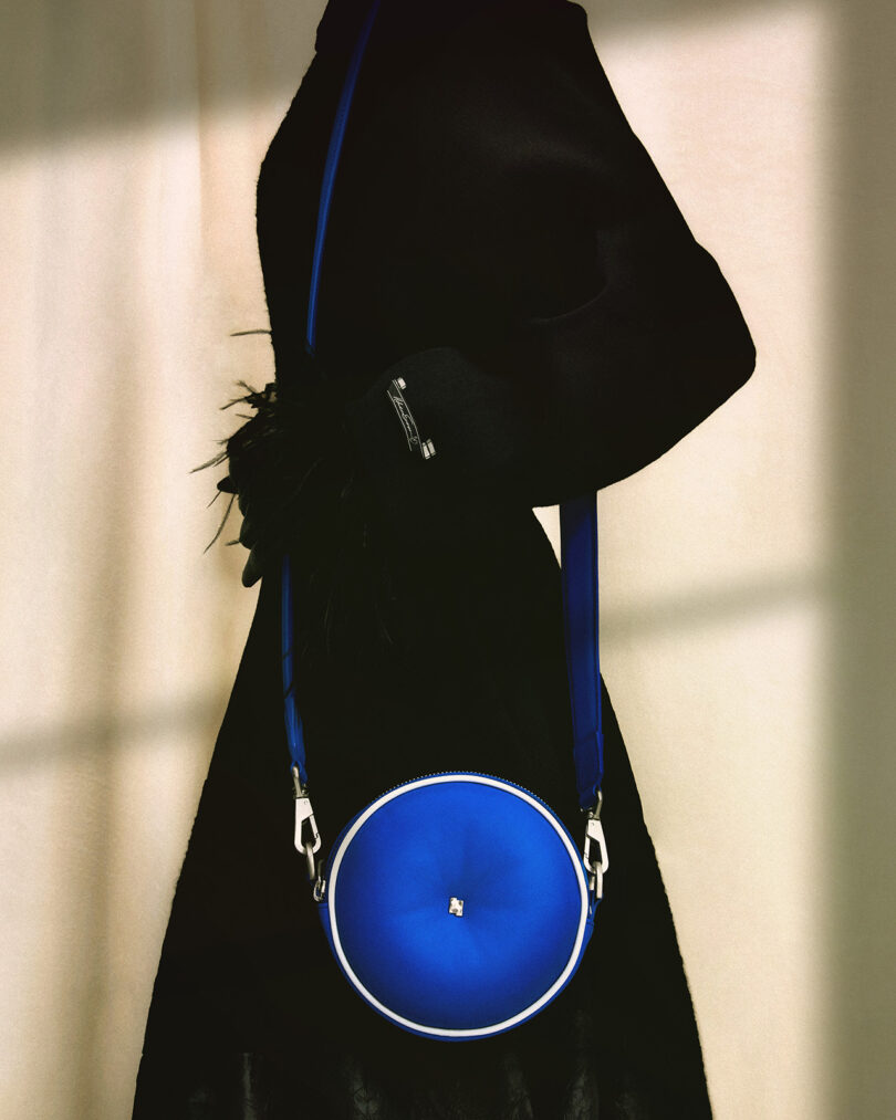 A blue circular Bluetooth speaker carrying bag with a strap draped over a person wearing an all-black garment, viewed from the side