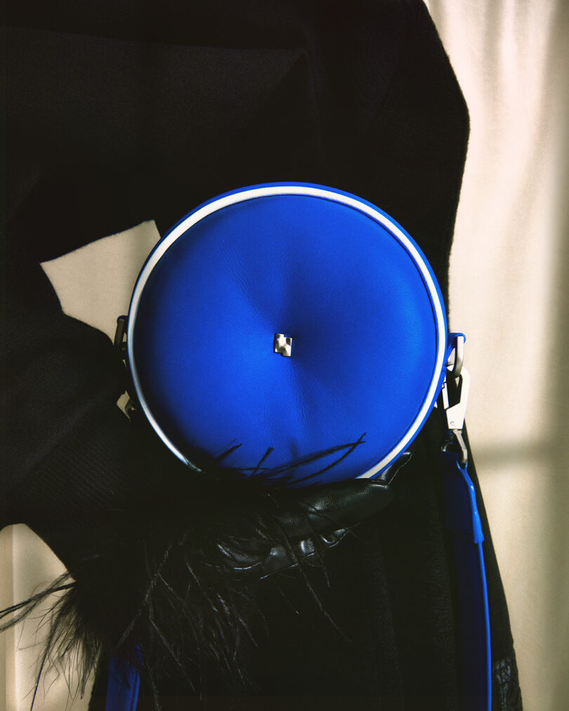 Blue round shoulder bag with a Bang & Olufsen's Beosound A1 ADER ERROR Edition Bluetooth speaker within being held next to the waist of a person dressed in all-black dress with feather detailing across the wrist.