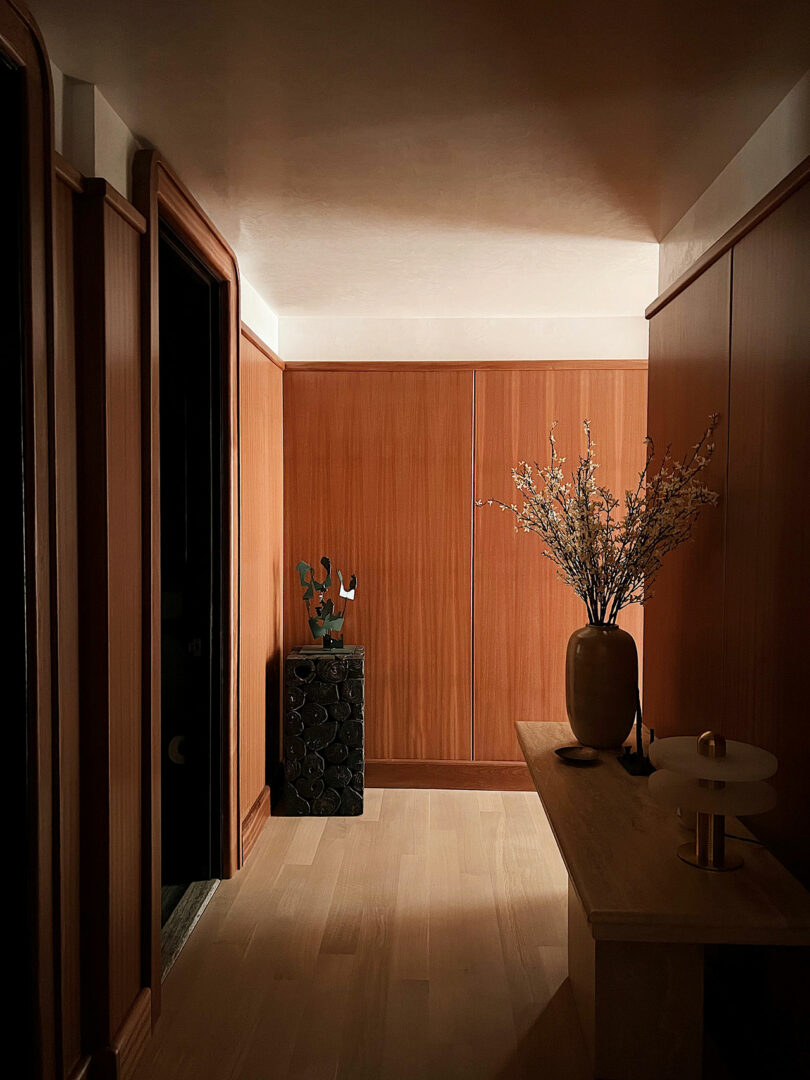 A dimly lit hallway in featuring wooden paneled walls, a vase with branches on a sideboard, and minimalistic decor.