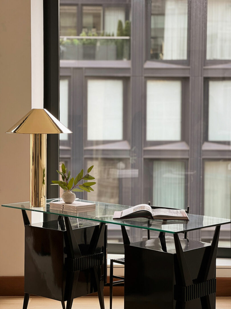Modern study area with a glass table, black chairs, open book, lamp, and a small plant, overlooking large windows with city view.