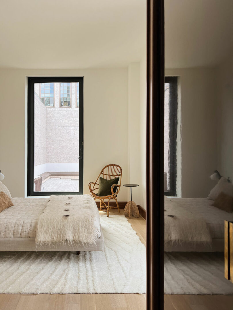 A minimalist bedroom with two beds, a large mirror, and a chair by a tall window overlooking a city building.