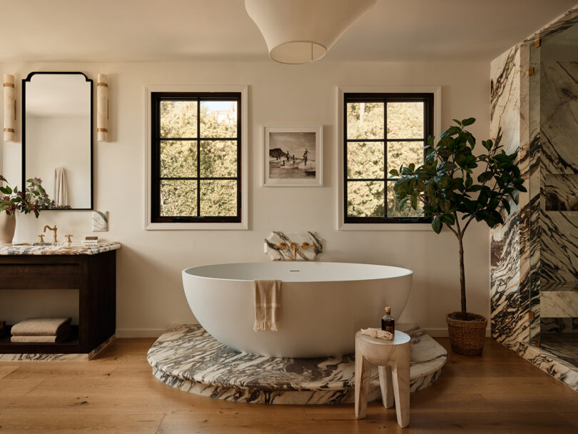 Elegant bathroom with a freestanding tub, marble floors, and a small wooden table, flanked by greenery and framed by multiple windows.