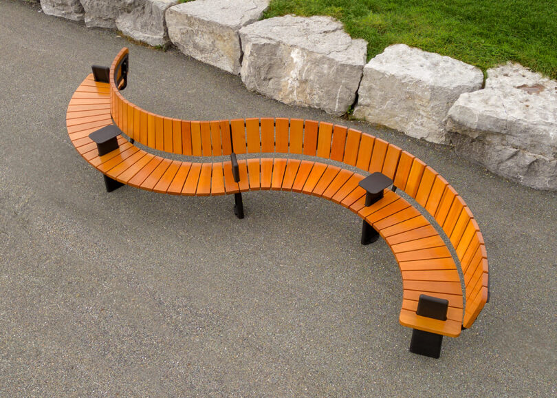 A curved wooden wooden bench with black metal supports, positioned on a concrete path beside a grassy area and stone wall.