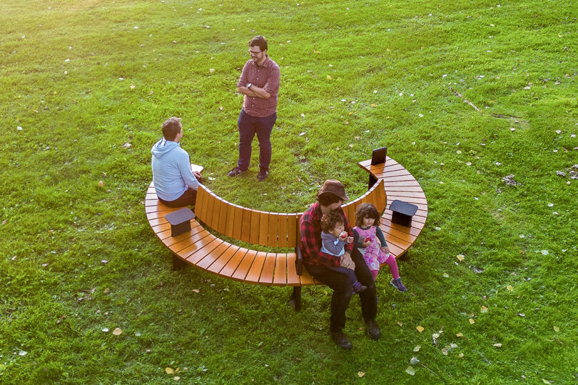 People conversing on a circular wooden bench surrounded by a grassy area.
