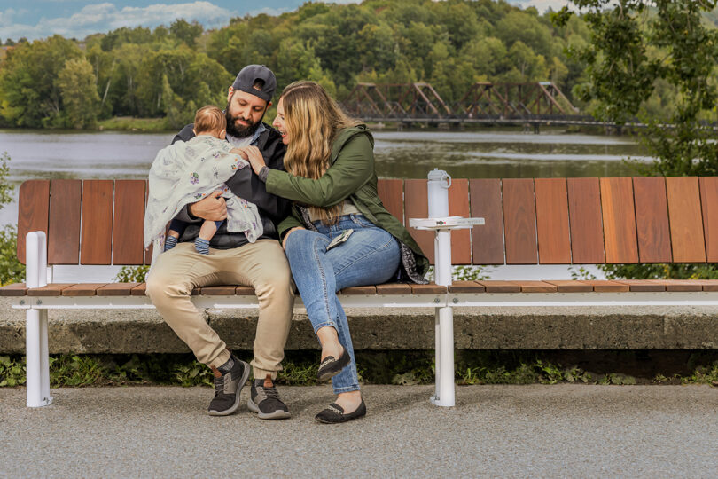 A family of three on a park bench by a river, with a bridge in the background.