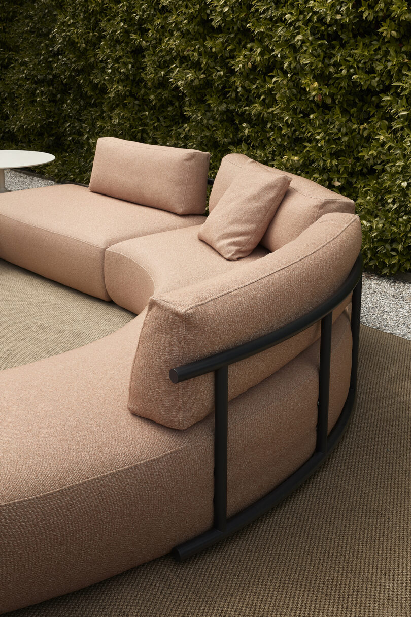 Modern outdoor lounge area with pink curved sofas on a rug against a dense hedge backdrop.