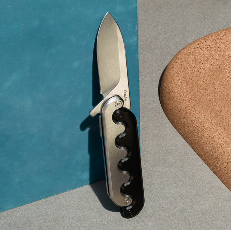 A Sidewinder pocket knife with an 2-tone interlocking hilt handle with its blade extended out against a blue and grey background.