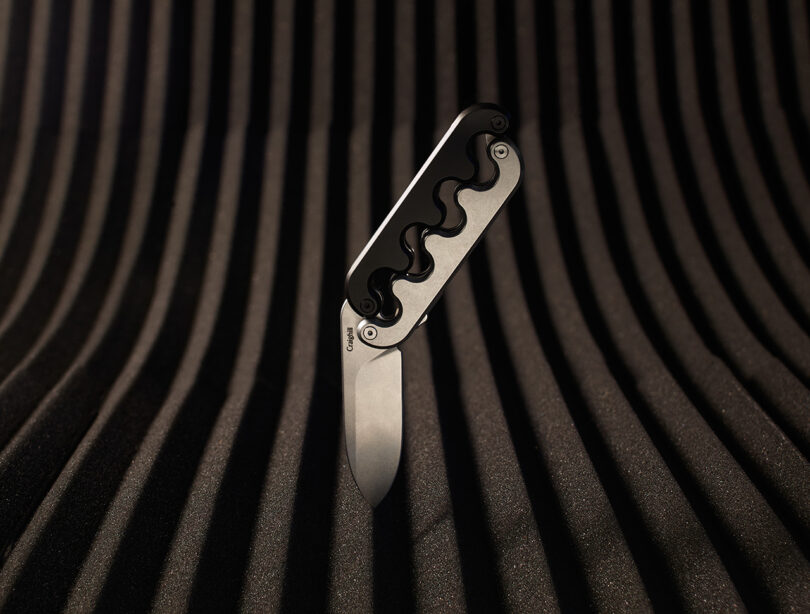 Sidewinder pocket knife with a 2-tone interlocking hilt handle set blade down across a striped/vented surface.