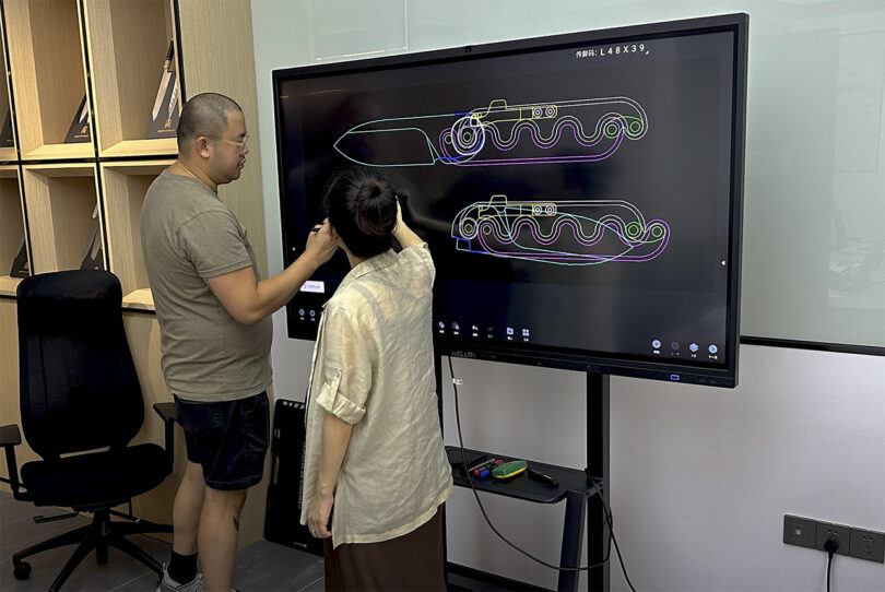 Two people discussing a digital rendering of the Sidewinder pocket knife with an 2-tone interlocking hilt handle design on a large screen, one holding a Sidewinder pocket knife.