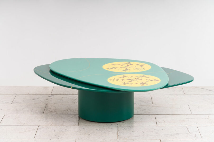 A modern coffee table designed by Djivan Schapira with a round, green top and three yellow circular inserts featuring artistic designs, set on a single cylindrical leg against a white backdrop.