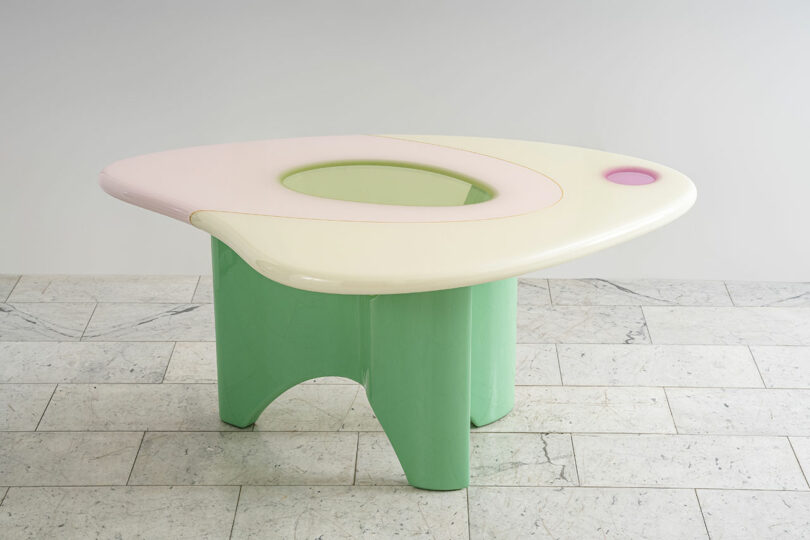 A modern, three-legged table with a green base and a uniquely shaped pastel pink and cream top, featuring a circular cutout crafted by Djivan Schapira, set against a plain grey background