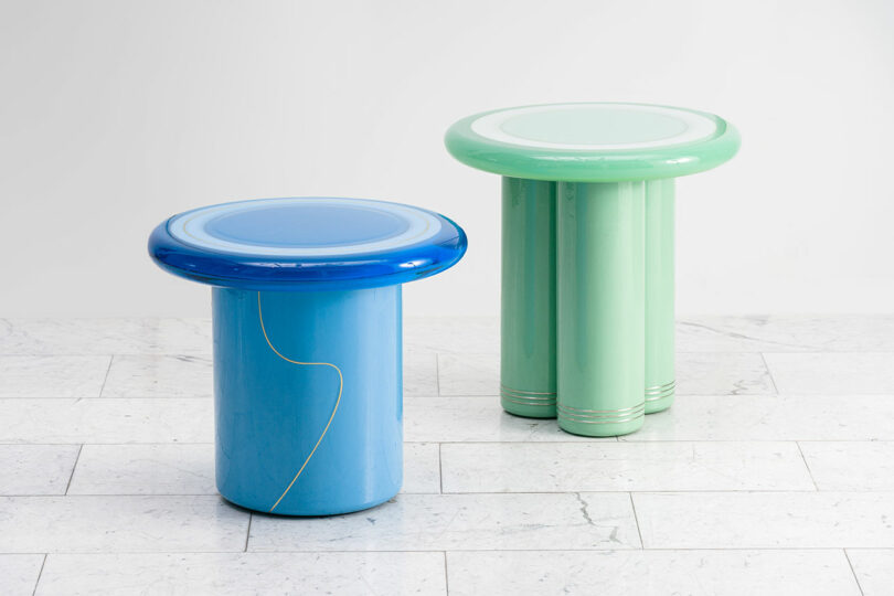 Two cylindrical stools designed by Djivan Schapira, one green and one blue, on a white floor against a white wall, with the blue stool showing imbedded bronze line-work.