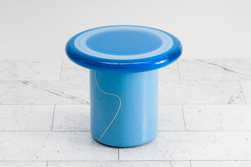 A blue cylindrical stool with a glossy finish, featuring a thin yellow line design by Djivan Schapira, on a marbled tile floor.