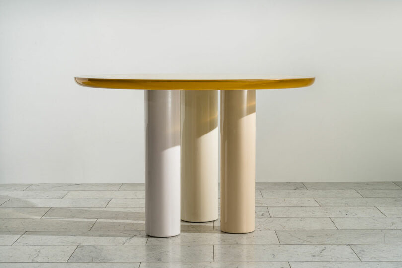 A modern table designed by Djivan Schapira with a glossy golden top and three cylindrical white legs, positioned on a tiled floor against a plain white wall.
