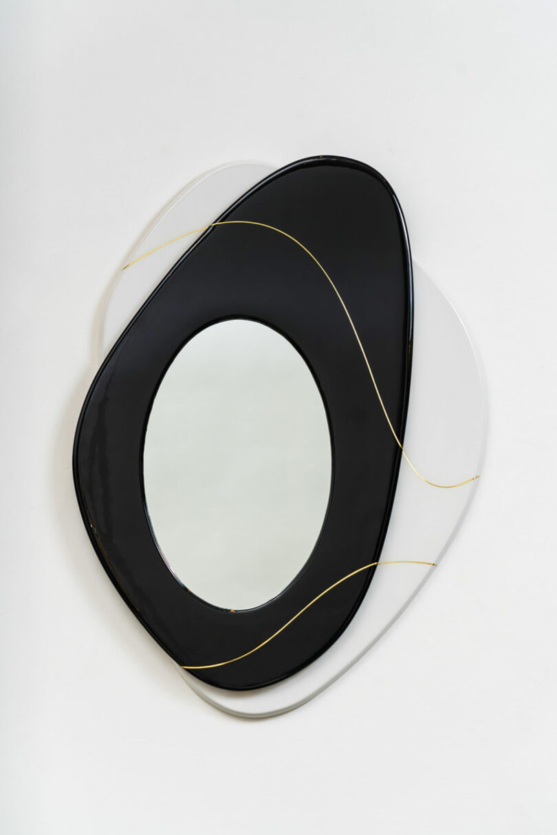 Modern wall mirror featuring an oval shape with layered black and white frames, accented with delicate gold lines designed by Djivan Schapira, against a white background.