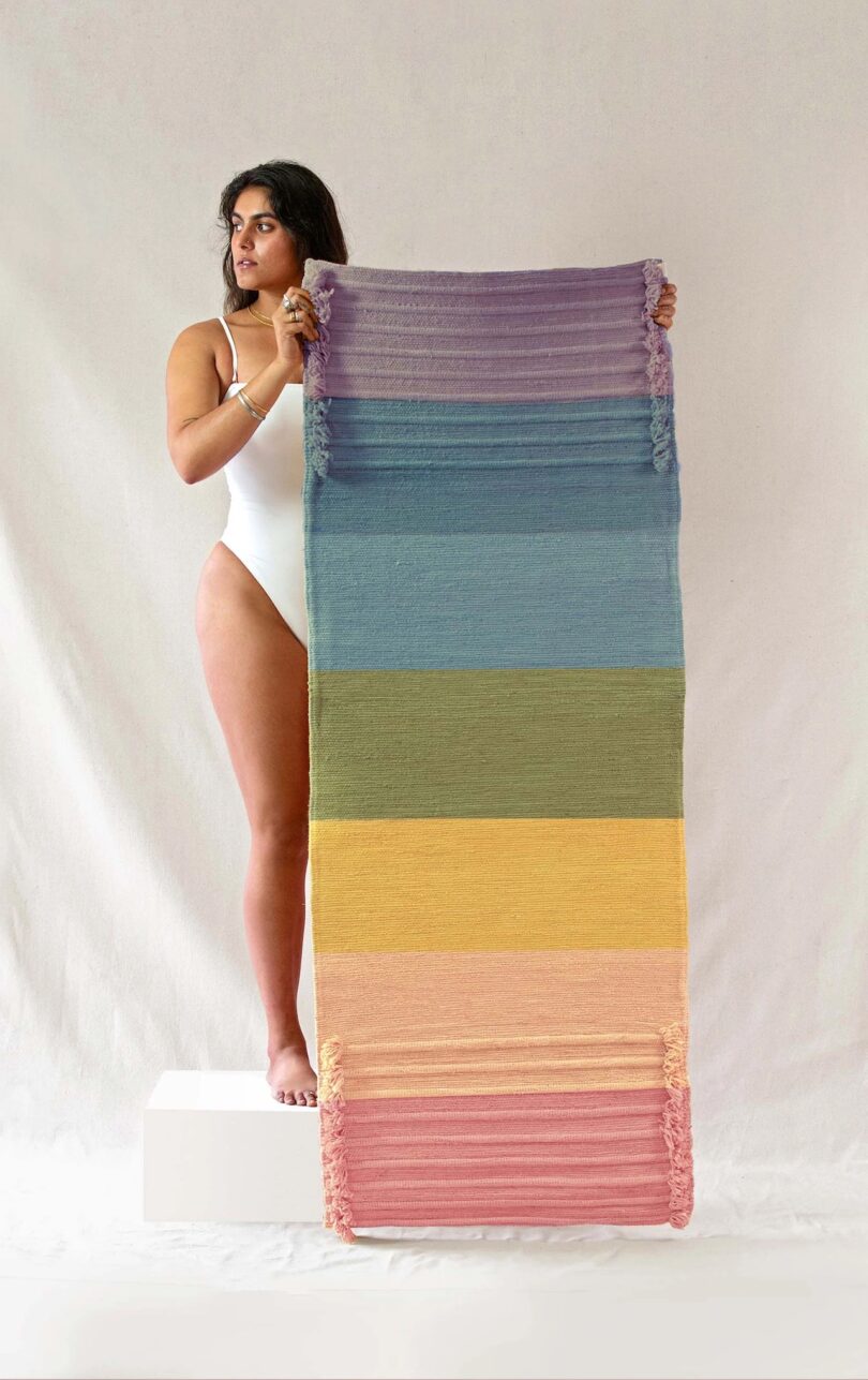 a woman is holding up an Oko Living Chakra Energy - Herbal Yoga Mat; from top to bottom, the mat is purple, blue, light blue, green, yellow, peach/light orange, and dark pink