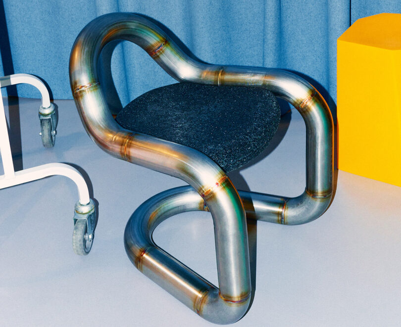 Metallic tubular chair with a continuous, flowing design and a black cushion, set against a blue backdrop.