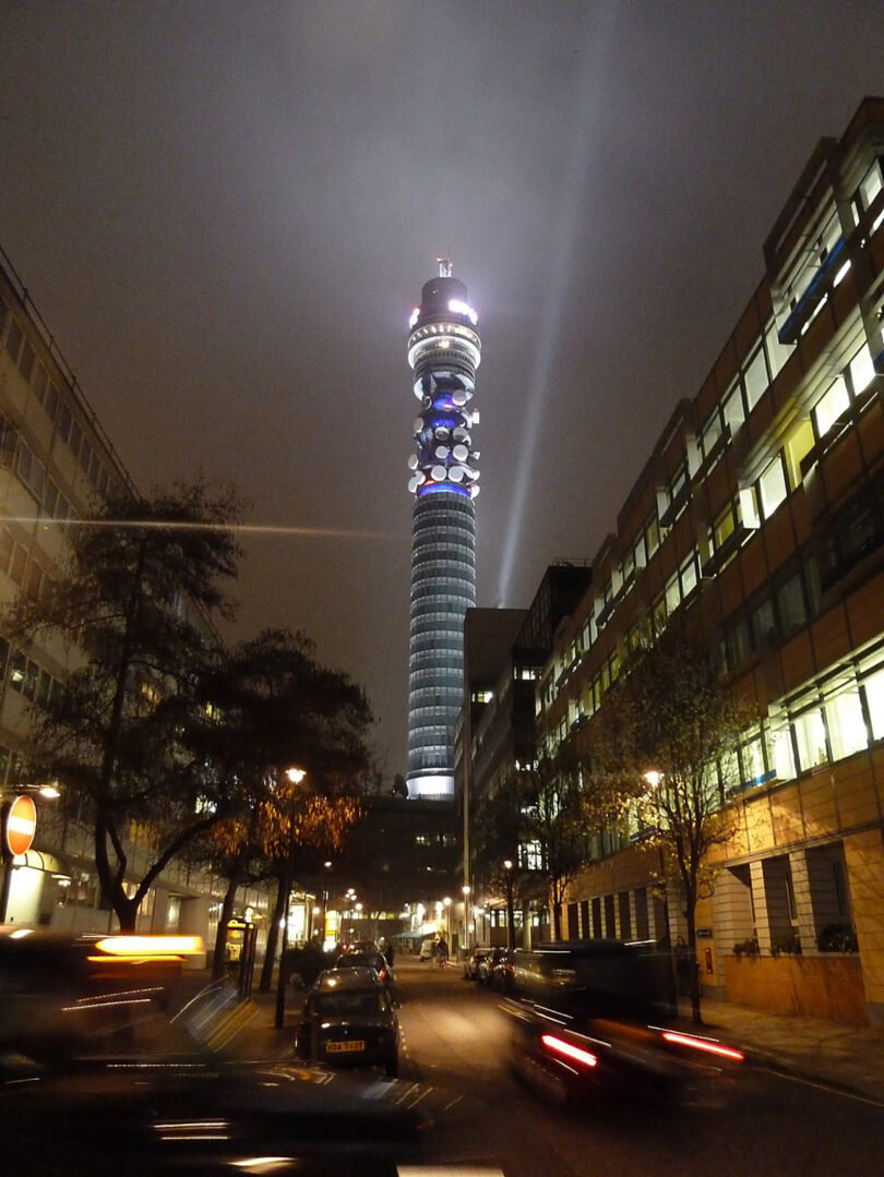 Night view of the bt tower in london, illuminated with colorful lights, seen from a nearby street with moving vehicles.