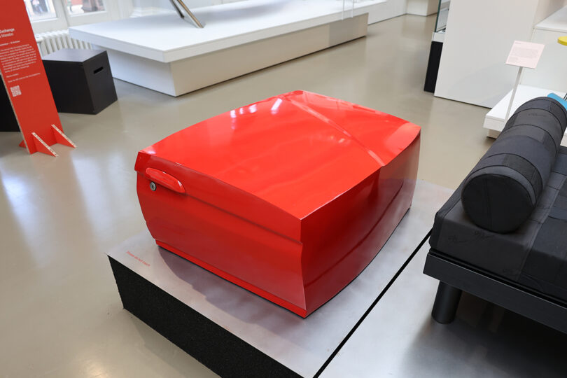 A shiny red, rectangular modern art installation by displayed on a platform in a gallery setting.